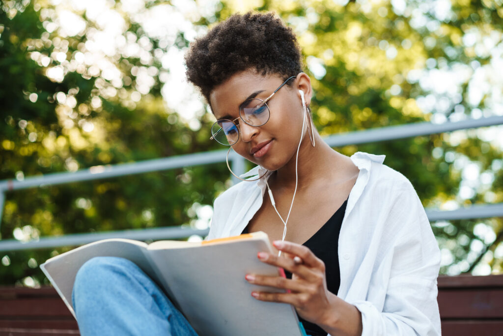 Photo of optimistic african woman sitting outdoors in park while listening music and reading book